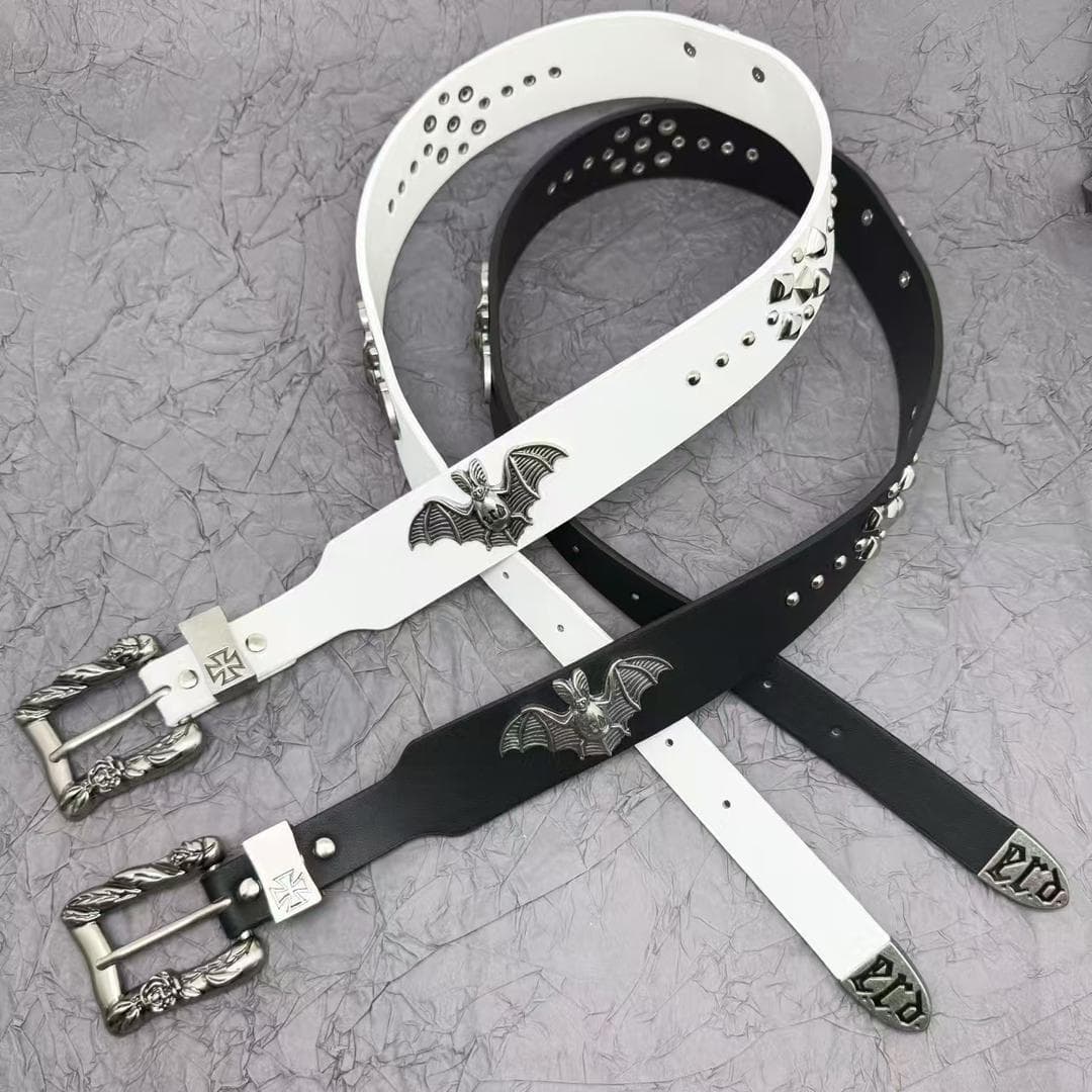 White and black bat emblem studded leather belts with ornate silver buckles on textured background.