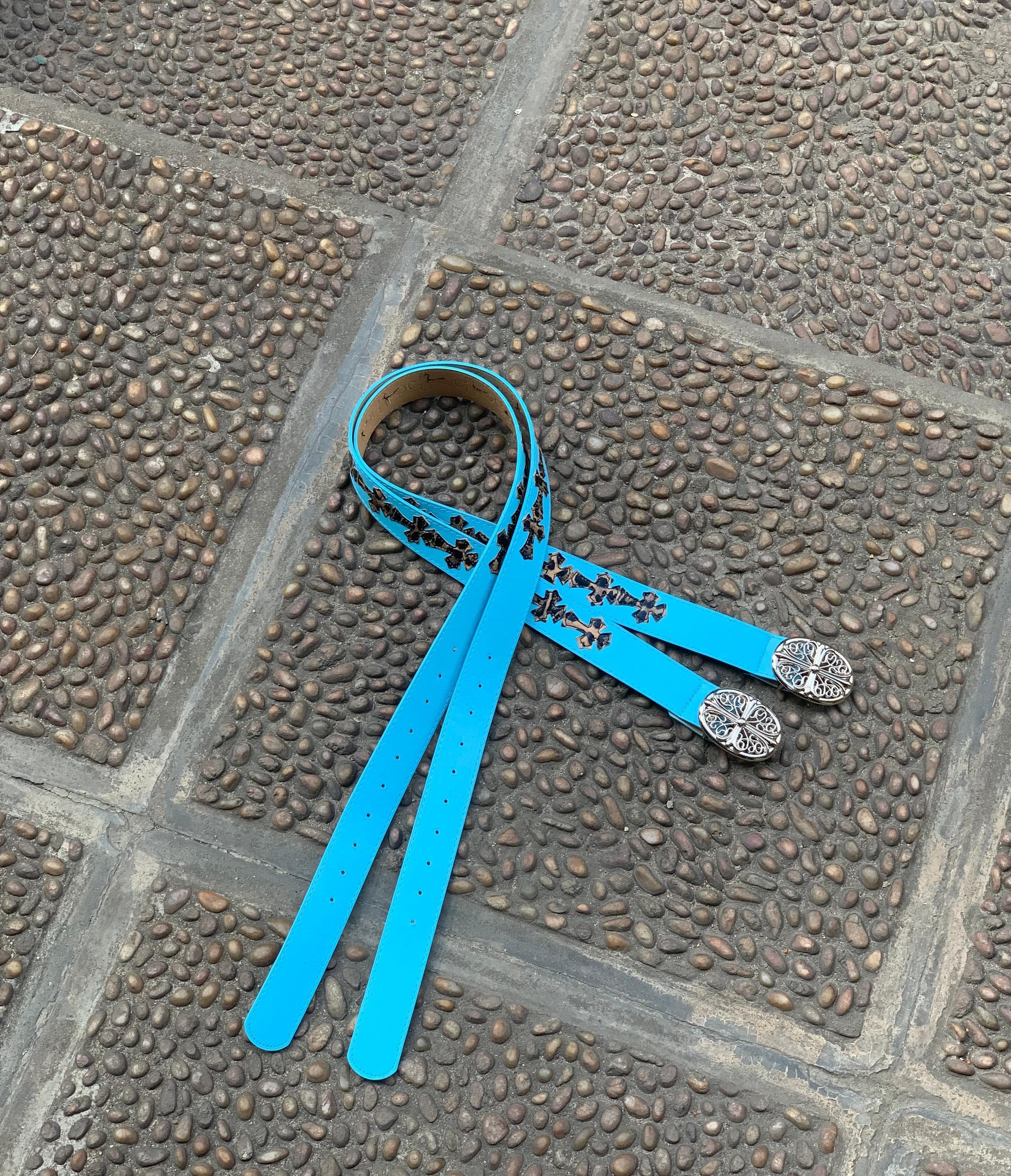 Bright blue leather belt with black cross patterns and two silver ornate buckles on cobblestone.