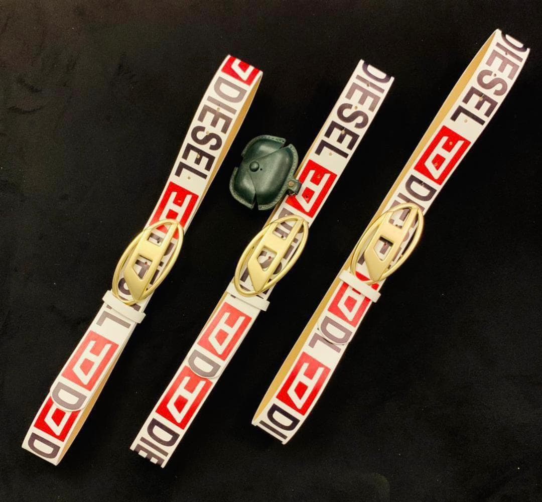 Three white Diesel logo belts with gold buckles displayed on black background alongside a small green pouch.