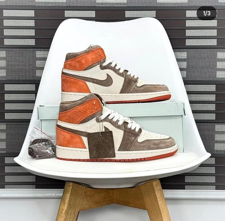 Nike Air Jordan 1 high-top sneakers in white, brown and orange color blocking displayed on a white chair with box and tags.