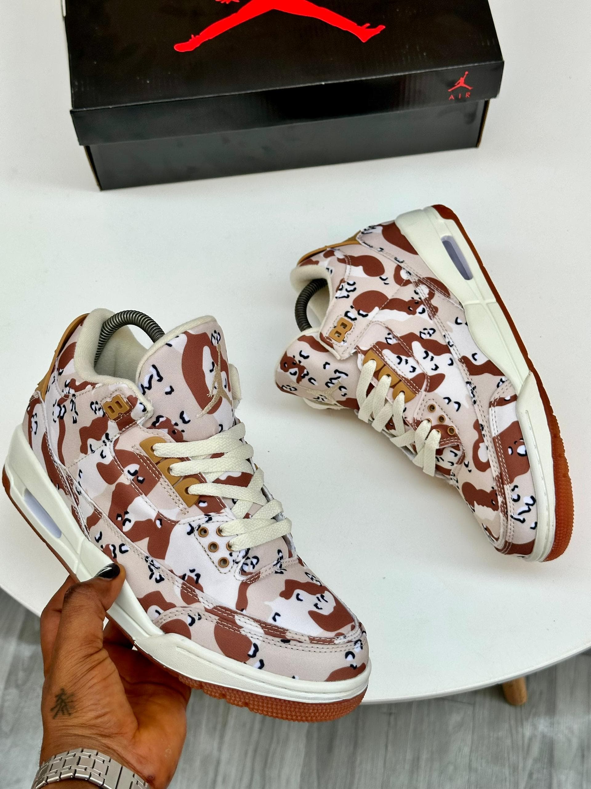 Camouflage print Air Jordan high top sneakers in brown and beige held in hand with box.