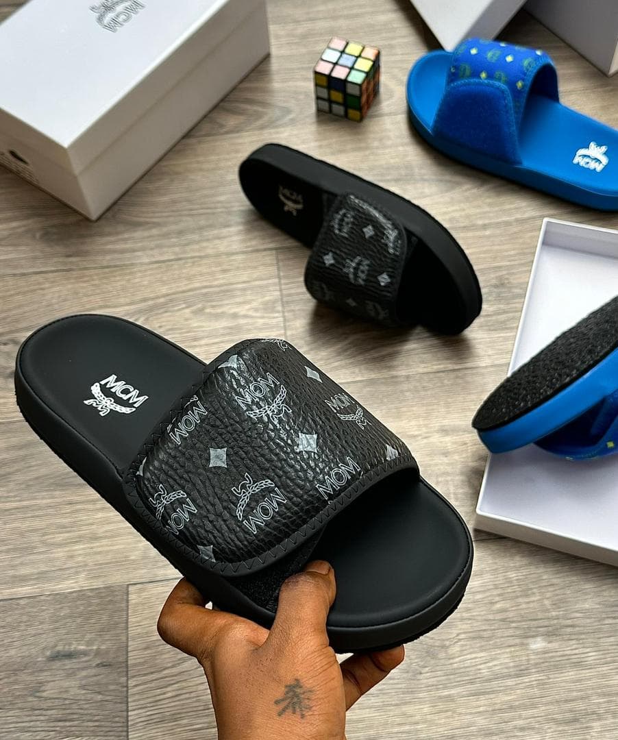 Black MCM monogram slide with white logos held in hand with other colorways visible.