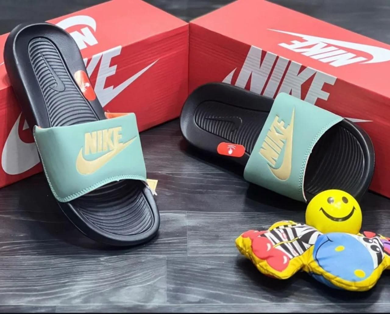 Teal Nike Victori One Slide with gold Swoosh next to red Nike boxes and smiley toy.