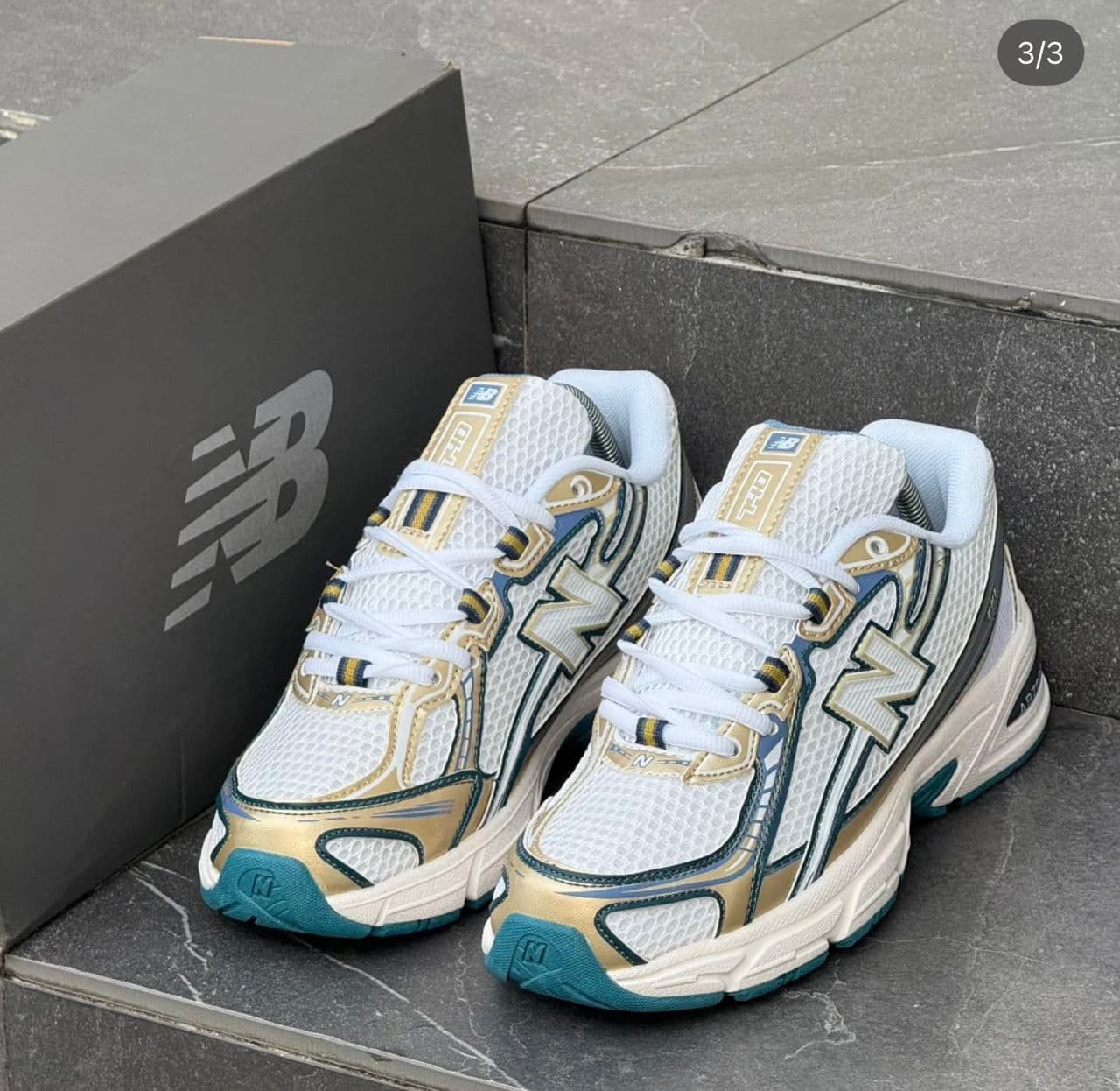 New Balance 9060 sneakers in white with gold and teal details next to NB box on steps.