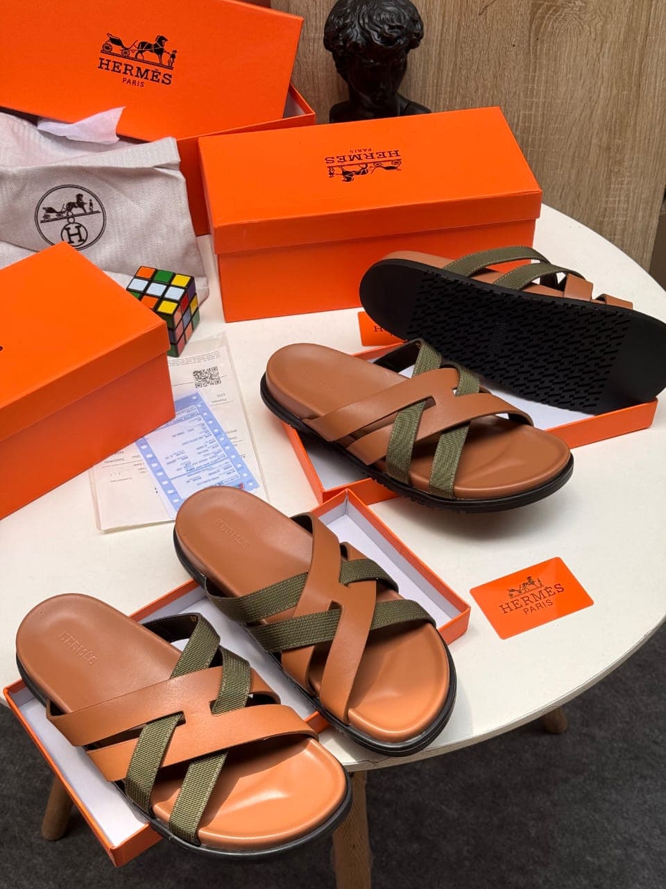 Brown and green strappy Hermes slides shown with boxes, Rubik's cube and accessories.