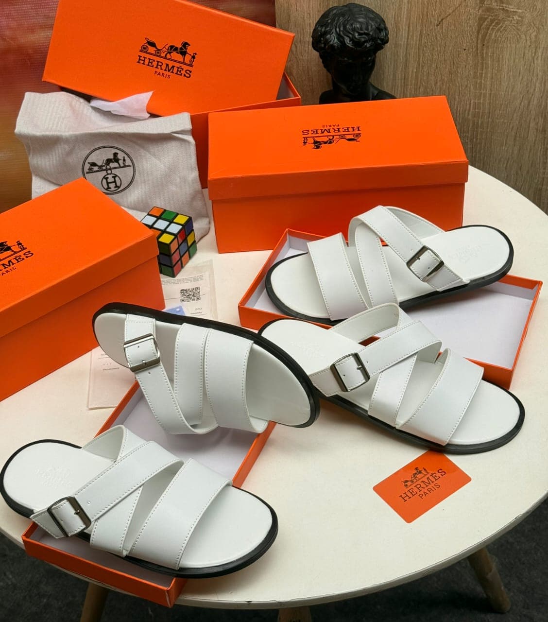 White Hermes multi-strap slides with buckle displayed with orange boxes and accessories.