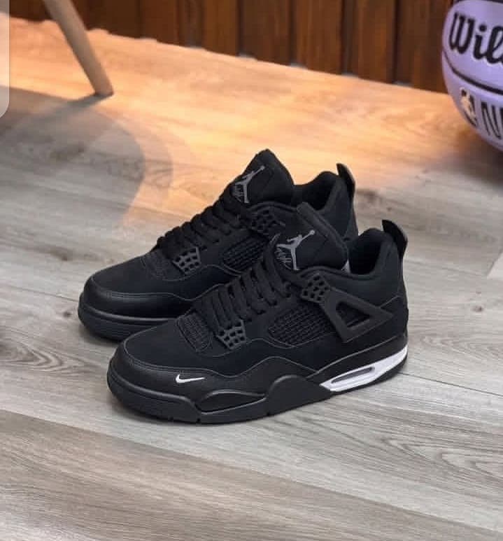 Pair of Nike Air Jordan 4 Black Cat sneakers placed on wooden floor with basketball in background.