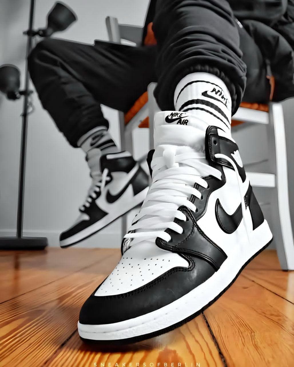 Black and white Air Jordan 1 High sneakers worn with black pants on wooden floor.