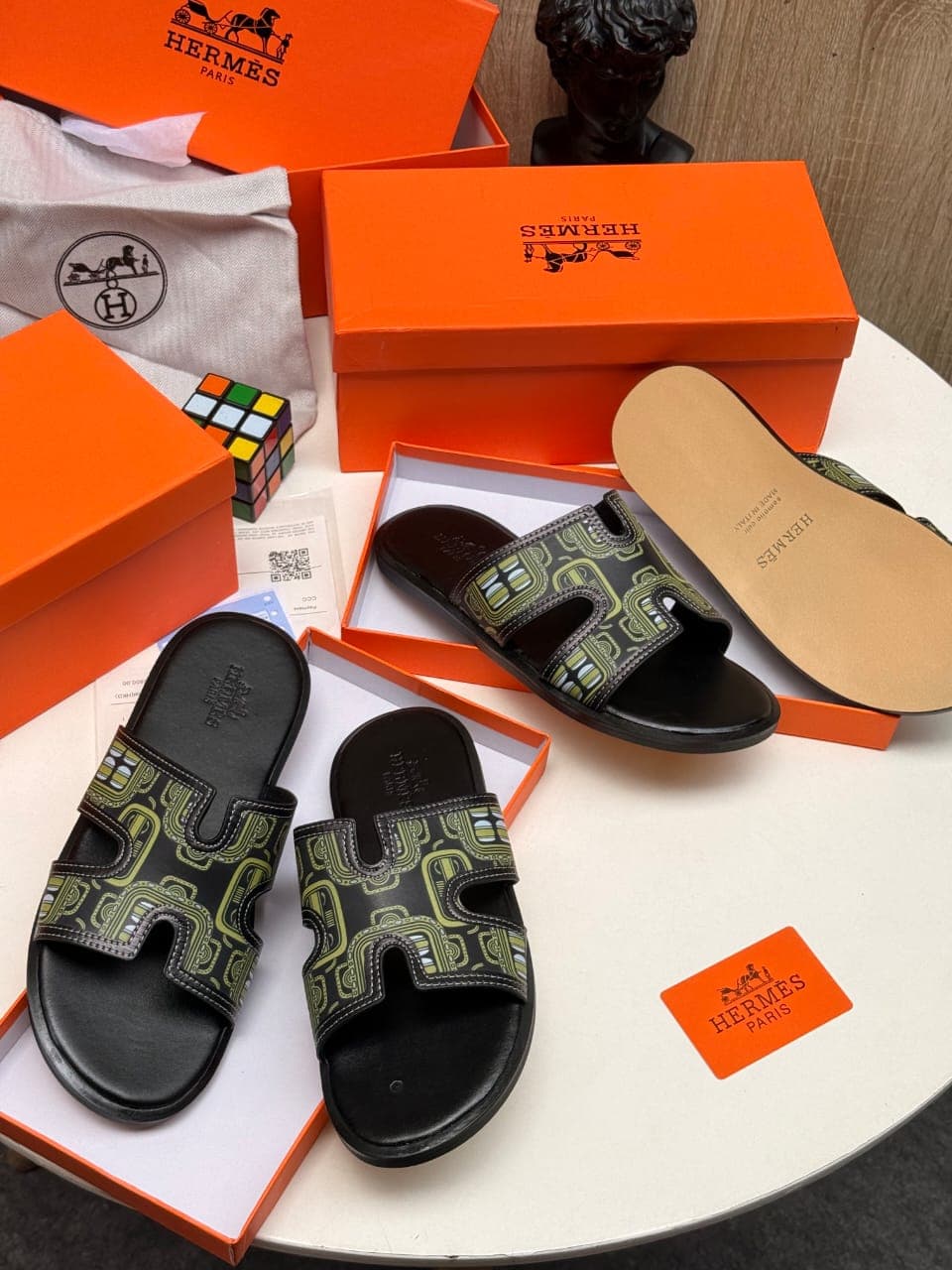 Black Hermes slides with green pattern on strap shown with orange boxes and dust bag.