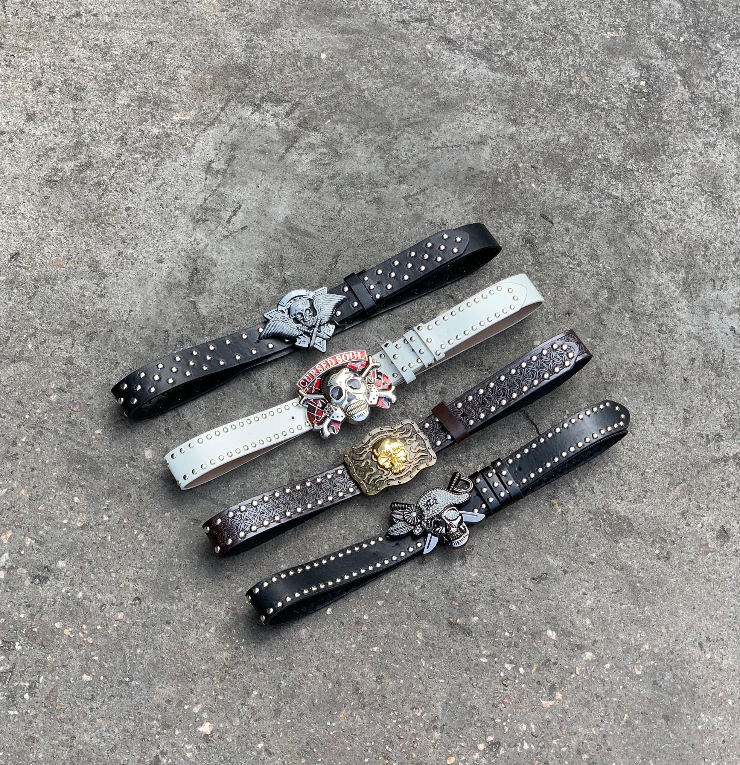 Four studded leather belts with skull and graphic buckles on gray surface.