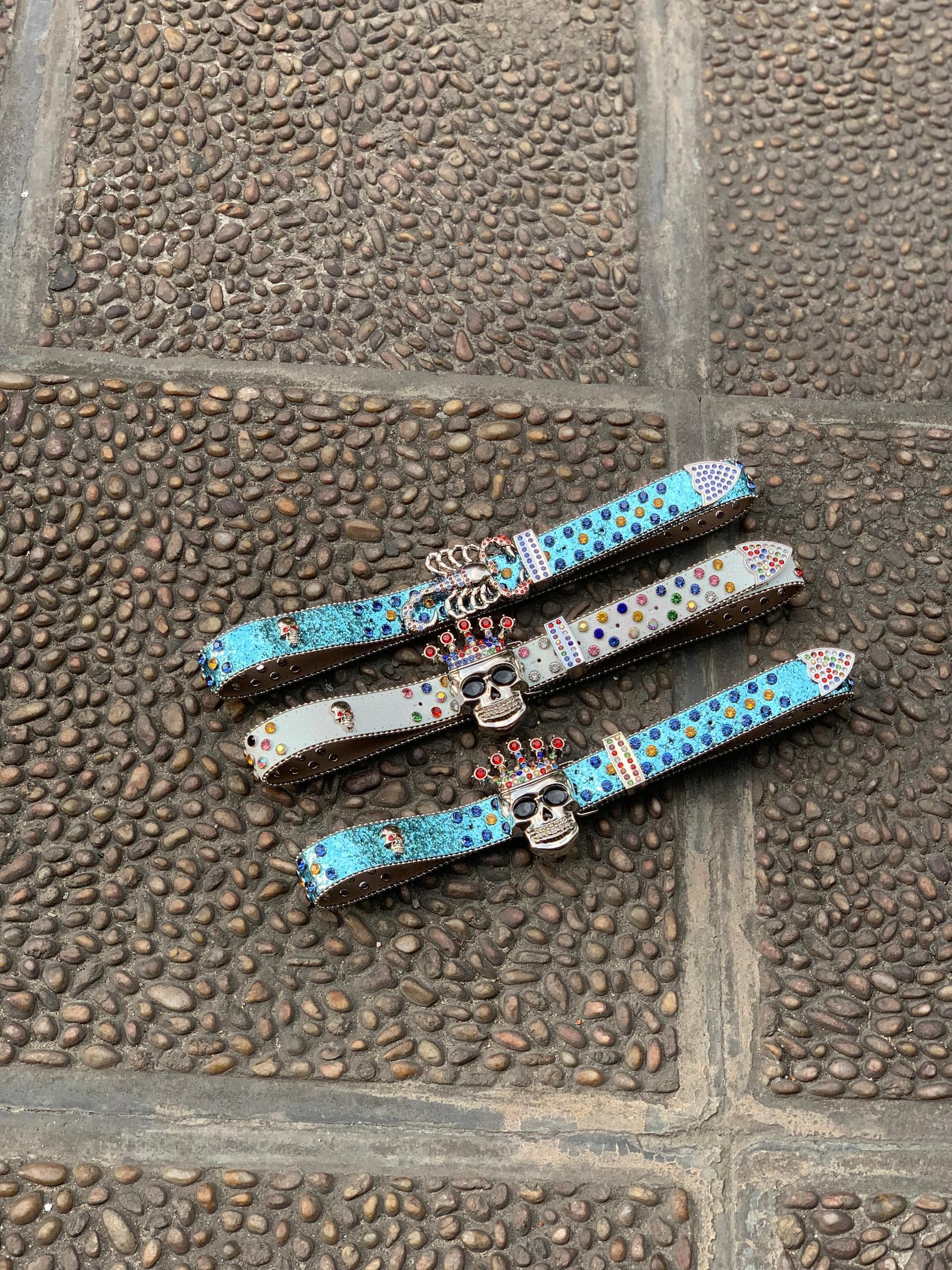 Assorted colorful rhinestone skull belts with crown buckles on stone pavement.