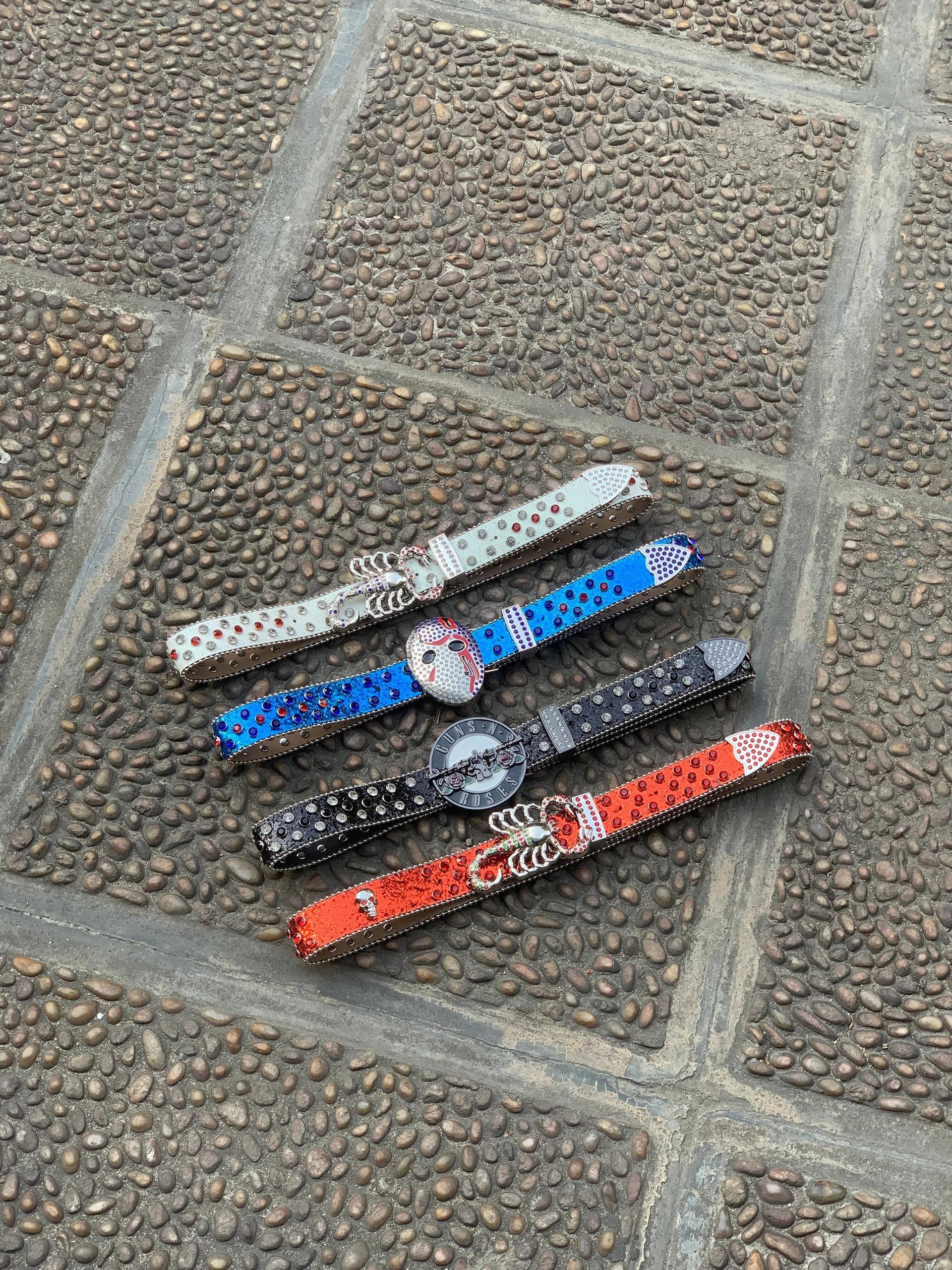 Four jeweled belts in white, blue, black and red with skull and Guns N Roses buckles on cobblestone.