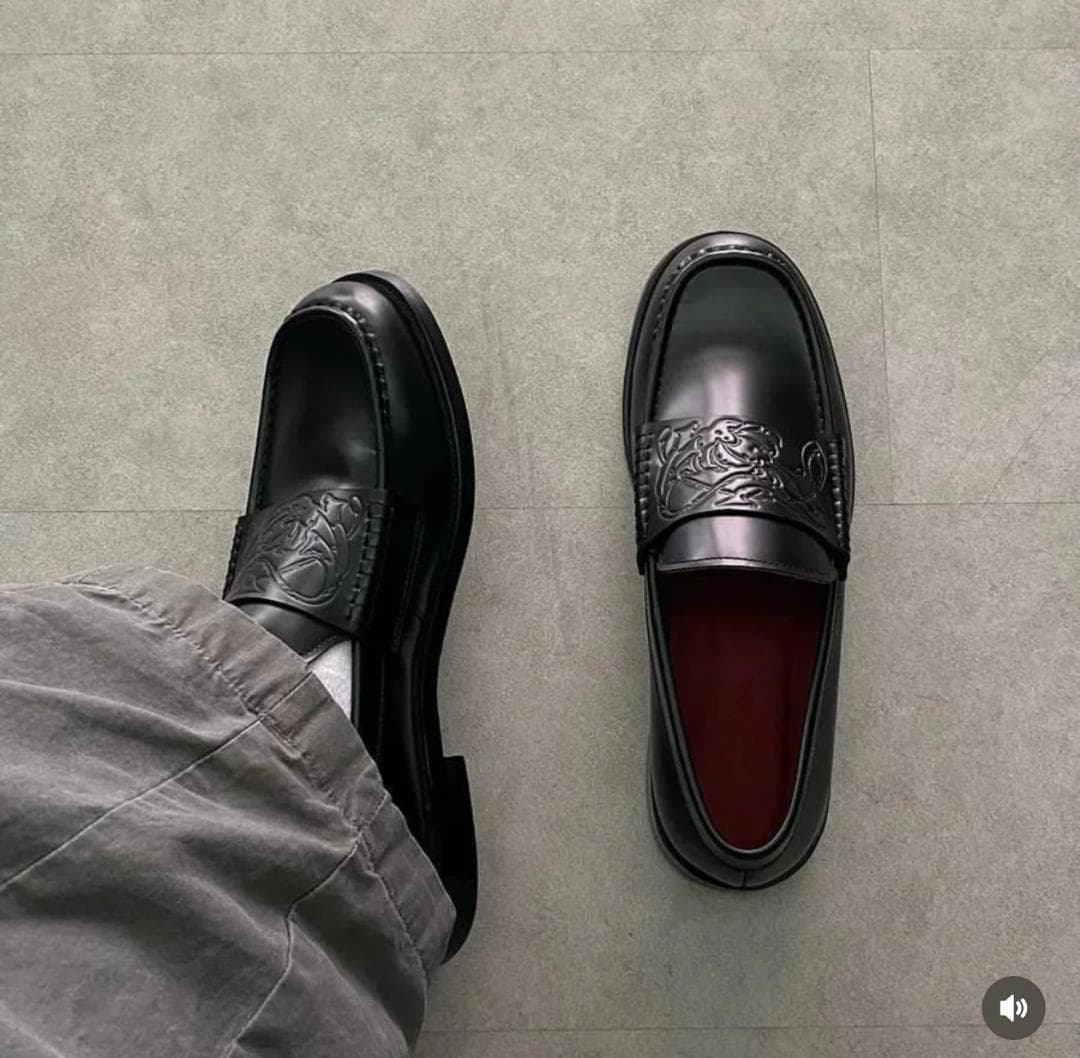 Black leather loafers with embossed design worn with grey trousers on a tiled floor.