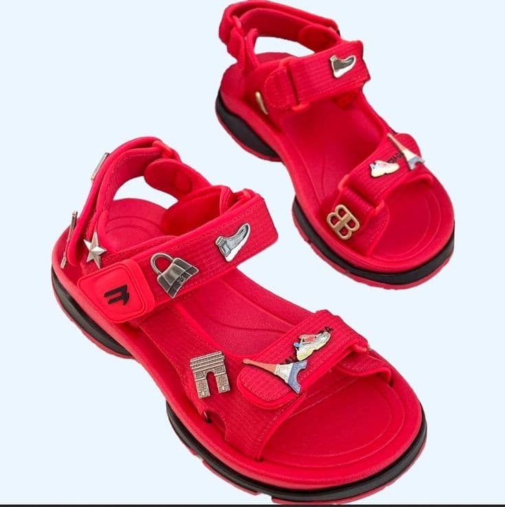 Pair of red sandals featuring multiple decorative metal charms on white background.