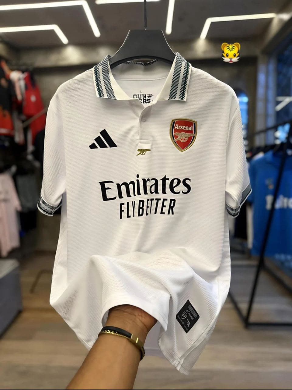 White Adidas Arsenal polo jersey held up on hanger in store.