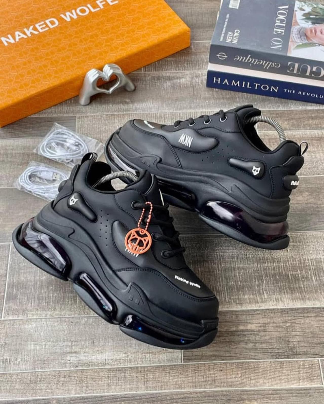 Black Naked Wolfe sneakers with orange charm and accessories on wood floor.