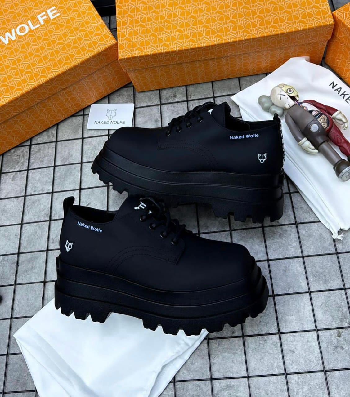 Two angles of black Naked Wolfe platform derby shoes on tiled floor with branding and accessories.