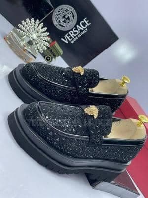 Pair of black rhinestone-covered platform loafers with gold emblems and Versace box in background.