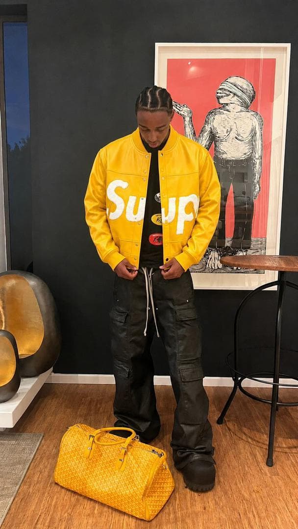 Man in yellow Supreme varsity jacket standing indoors beside a matching yellow bag.