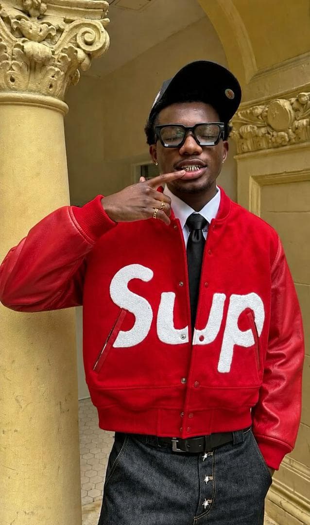 Man in red Supreme varsity jacket with white logo posing by a pillar.