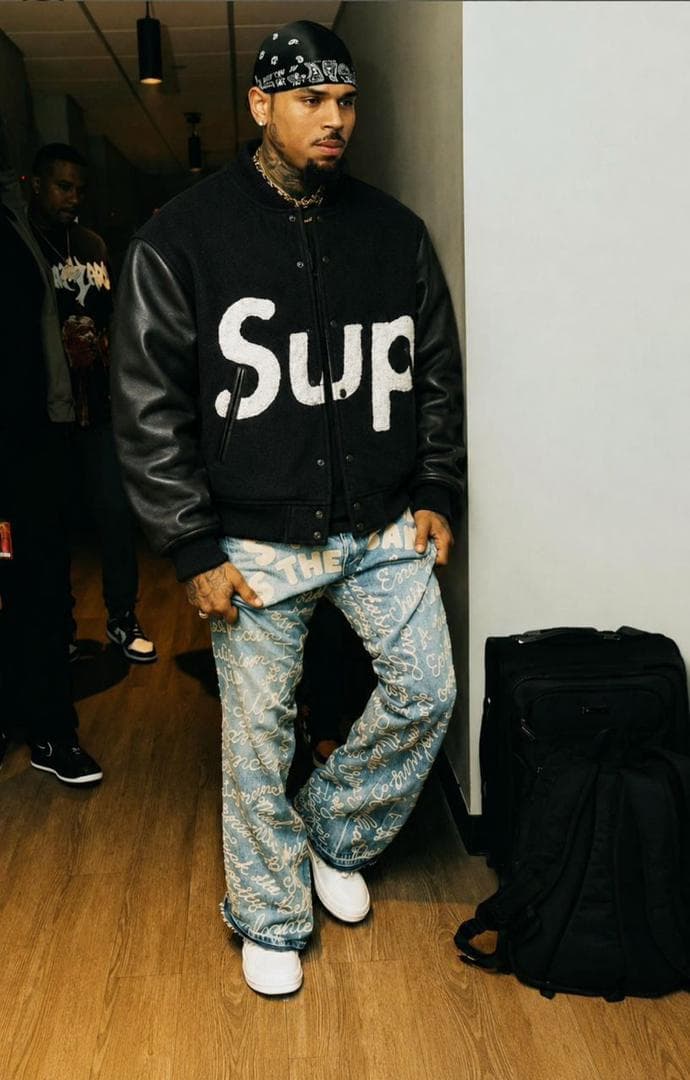 Man wearing black Supreme varsity jacket with white logo and patterned jeans.