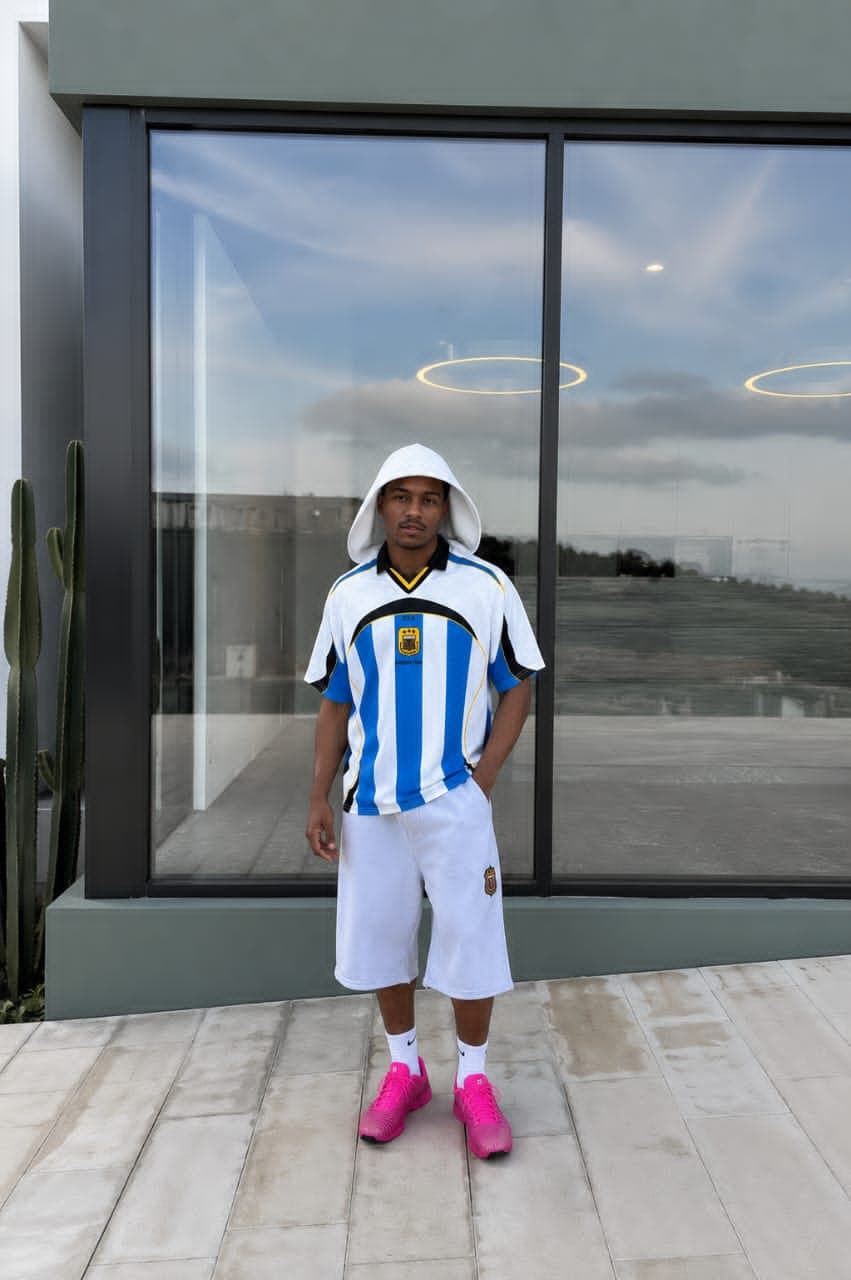 Man wearing Argentina striped jersey with white shorts, white hoodie, and pink sneakers standing outdoors.