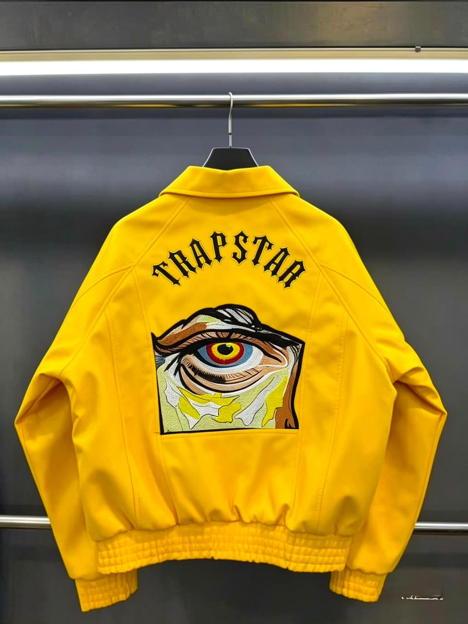 Yellow bomber jacket hanging with TRAPSTAR text and large colorful eye embroidery on back.