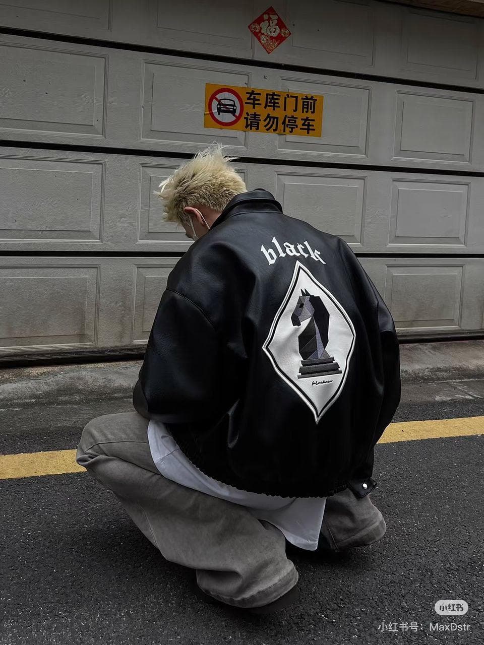 Person with blonde hair crouching in black bomber jacket showing 'black' text and chess knight graphic on back.