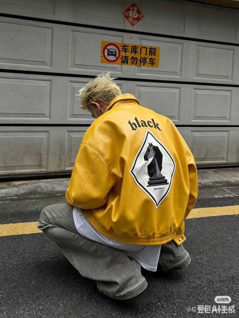 Person with blonde hair crouching in yellow bomber jacket showing 'black' text and chess knight graphic on back.