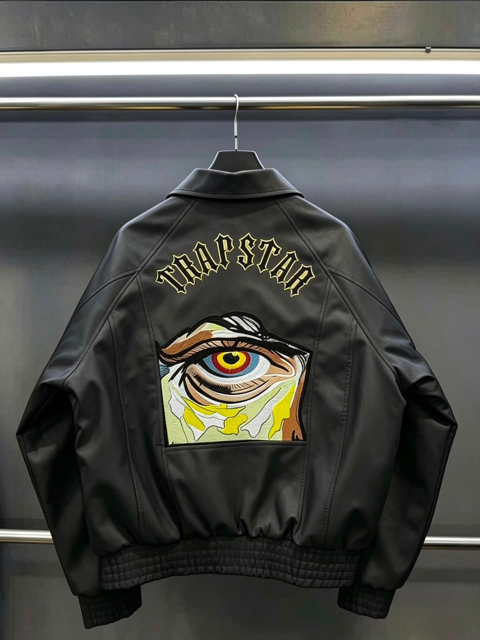 Black bomber jacket hanging with TRAPSTAR text and large colorful eye embroidery on back.