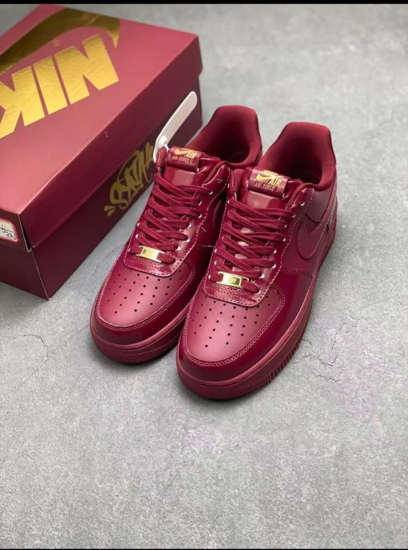 Pair of red patent Nike Air Force 1 sneakers with gold details beside red shoebox.