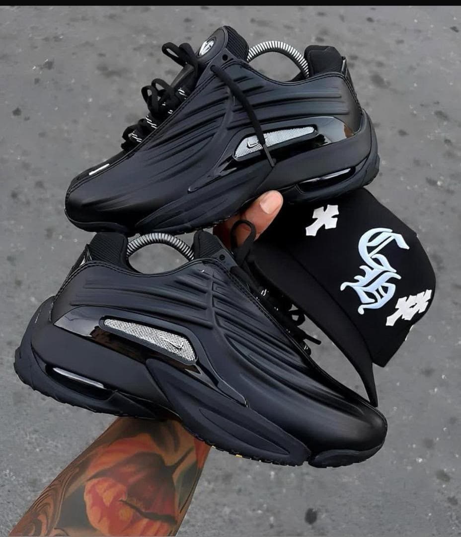 Pair of black Nike Air Max Plus sneakers held in hand with matching black cap.