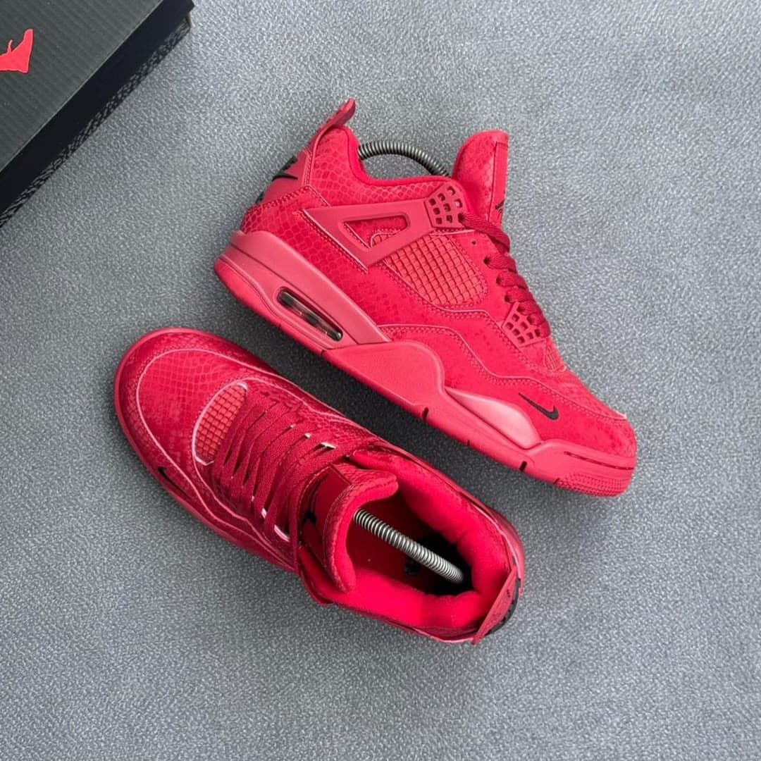 Pair of all-red Air Jordan 4 sneakers on gray surface with shoebox.