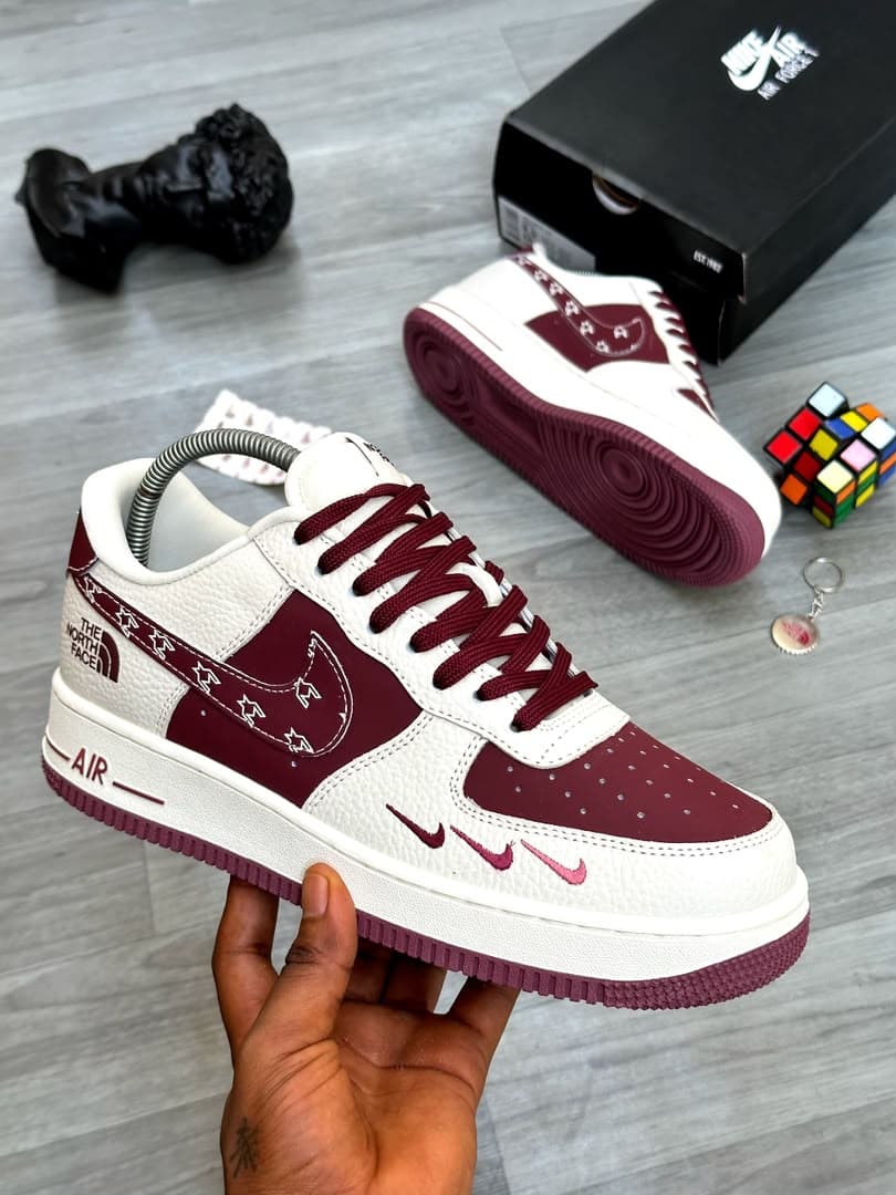 White and maroon Nike Air Force 1 Low with The North Face branding and star graphics held in hand.