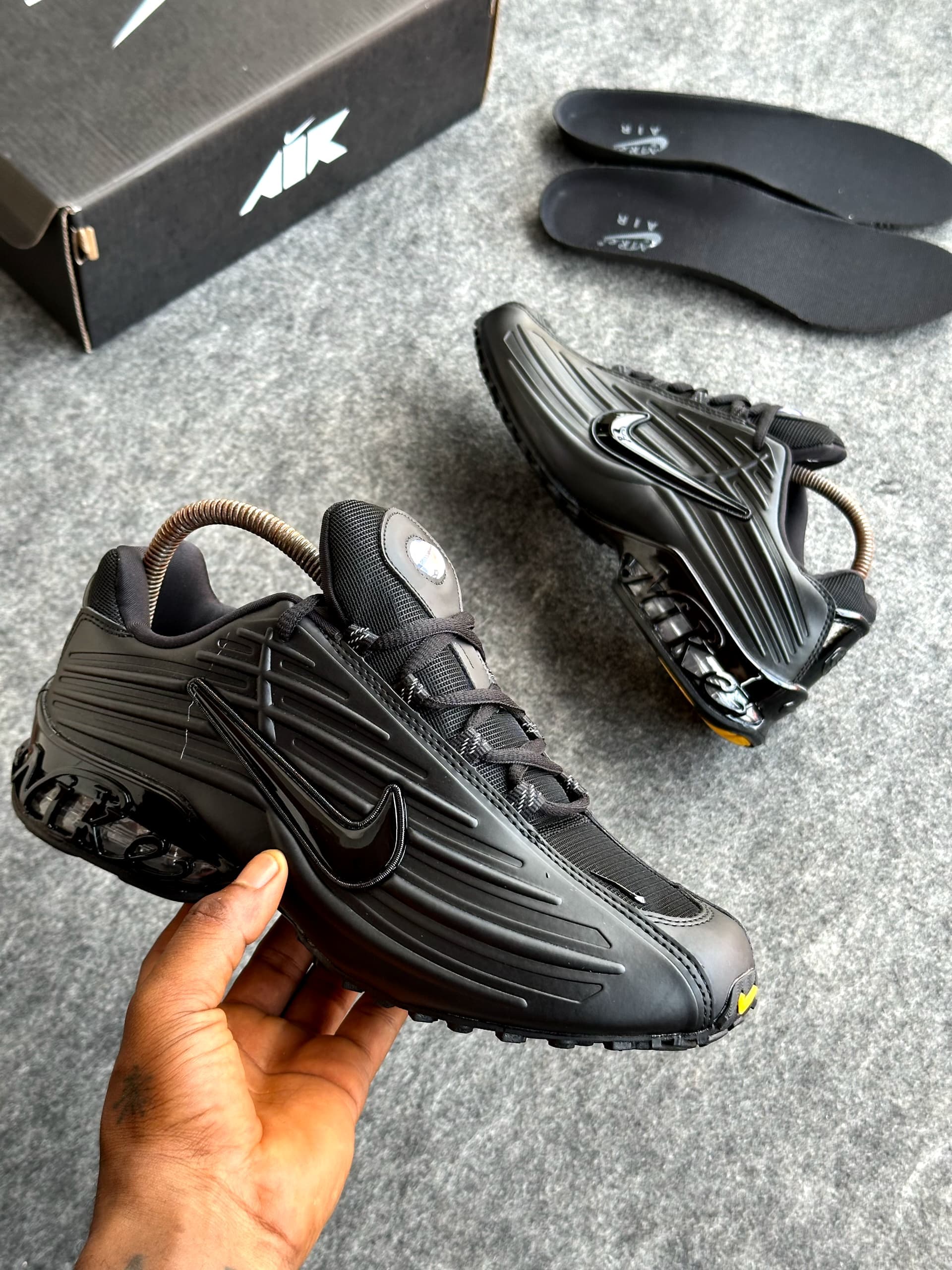 All-black Nike Air Max Plus sneakers held in hand with box and black insoles.