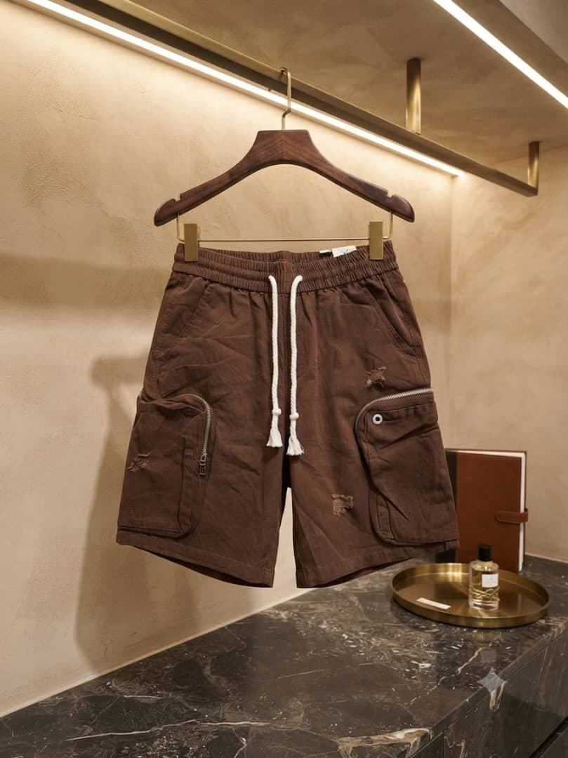 Brown unisex combat cargo shorts hanging on wooden hanger in modern interior.