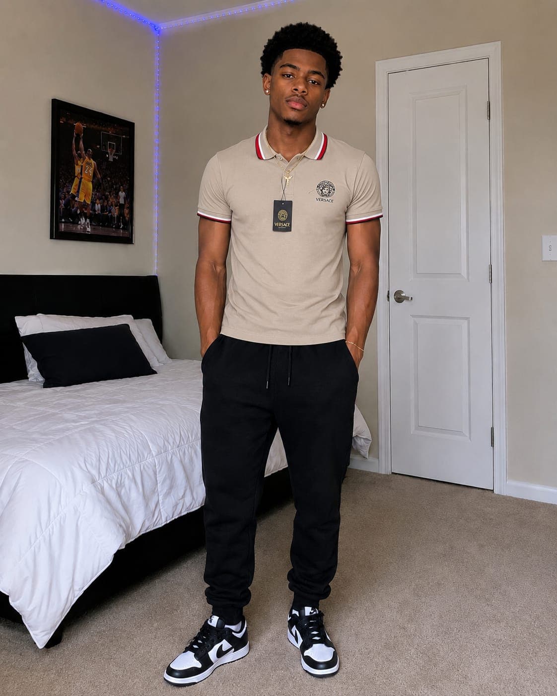 Man modeling beige Versace polo shirt with black joggers and sneakers in bedroom setting.