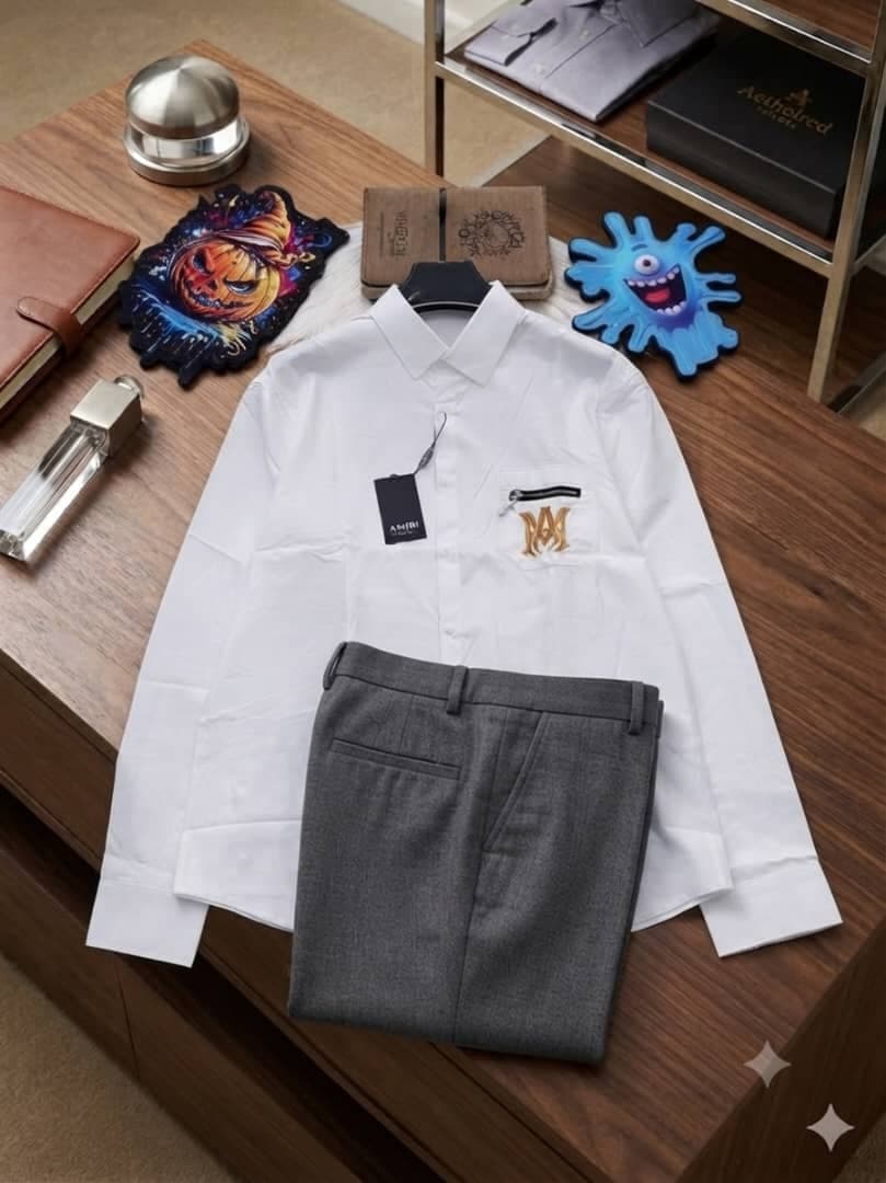 White Amiri MM logo shirt paired with gray trousers on wooden surface with props.