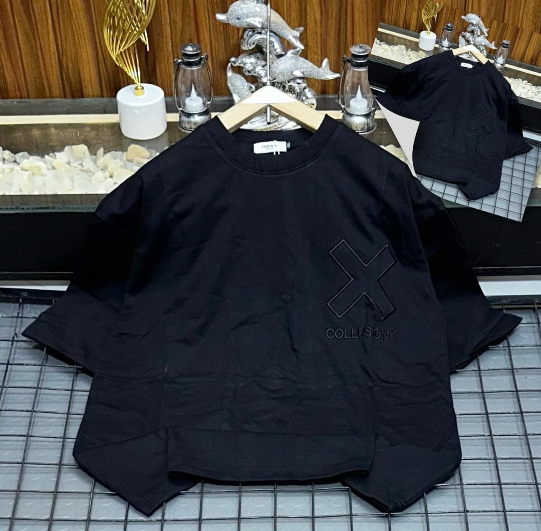 Black oversized t-shirt with large embroidered X and COLLUSION text laid on grid surface with inset view.