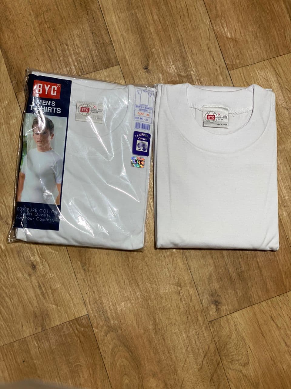 BYC men's white t-shirt in packaging beside unfolded version showing tags on wooden floor.