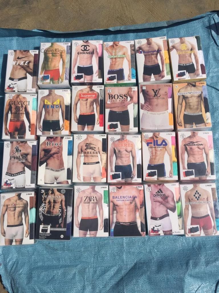 Assortment of over 20 different designer 3-pack boxer trunks laid out in rows on blue fabric, showing brands like Nike, Dior, Gucci, LV and more.