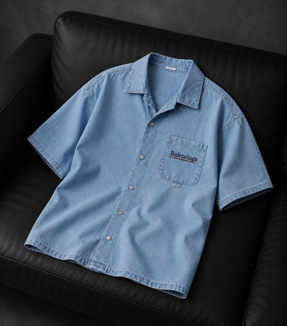 Light blue denim short-sleeve shirt with Balenciaga text on chest pocket, laid on black couch.