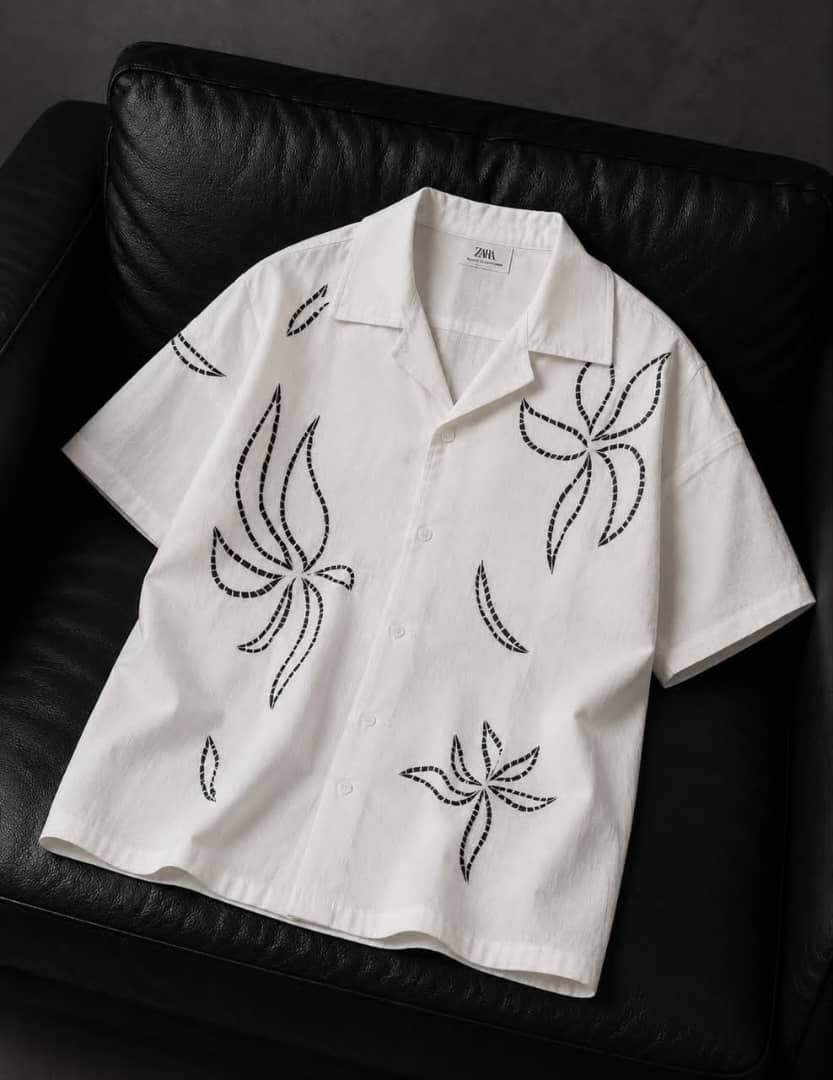 White short-sleeve button-up shirt with black embroidered floral patterns on black leather couch.