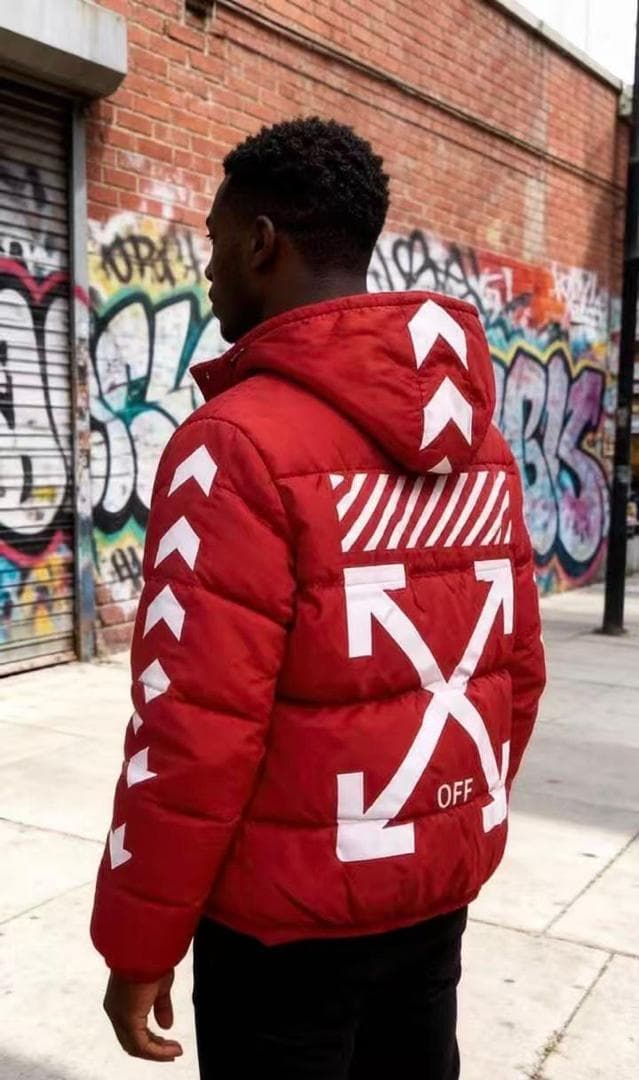 Man wearing red hooded puffer jacket with white arrow graphics on back and sleeves, standing against graffiti wall.