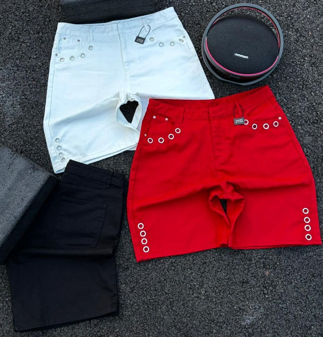 Flat lay of white, red, and black grommet denim shorts on asphalt.