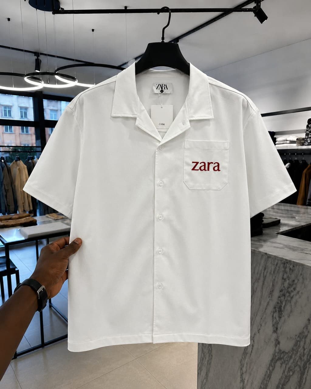 White short-sleeve button-up shirt with red 'zara' embroidery on chest pocket.