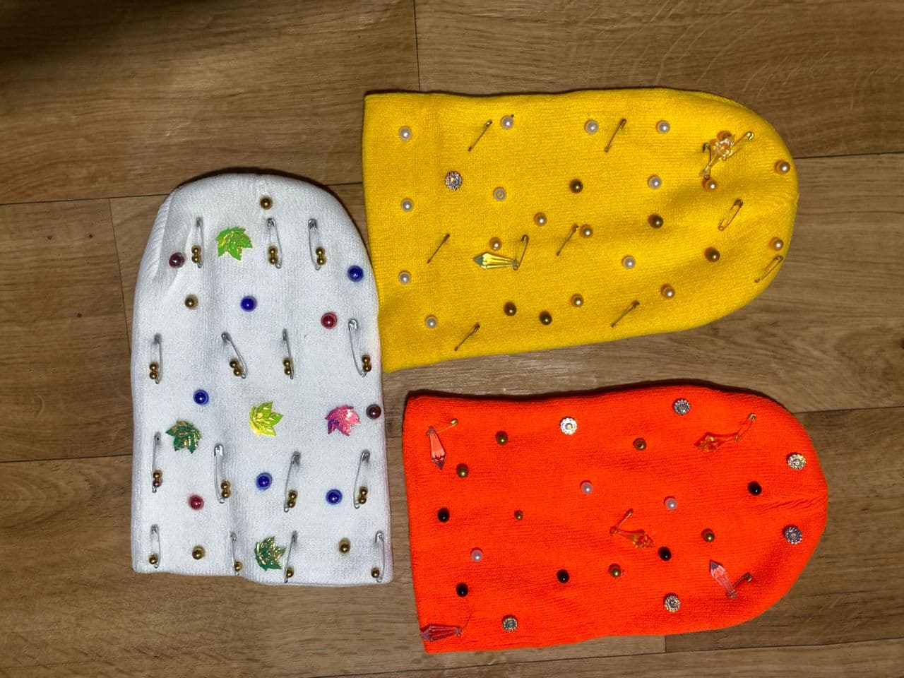 White, yellow, and orange knit beanies decorated with safety pins, beads, pearls, and charms on wooden floor.