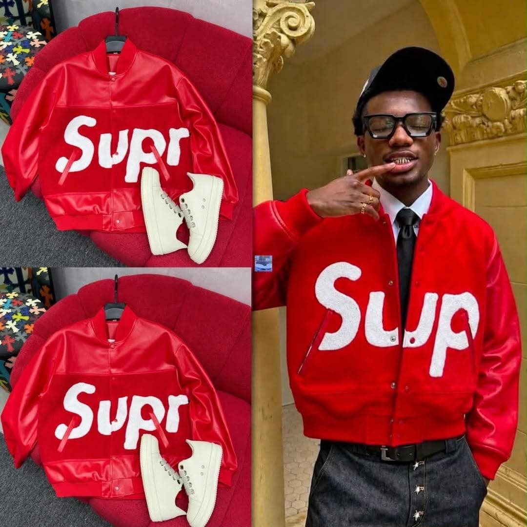 Red Supr varsity bomber jacket displayed on red surface with white sneakers and worn by model in glasses.