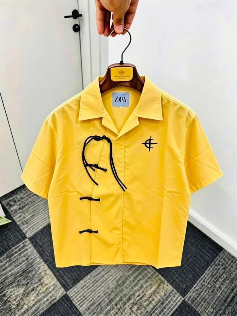 Yellow short sleeve shirt with frog buttons, drawstring and chest embroidery on wooden hanger.