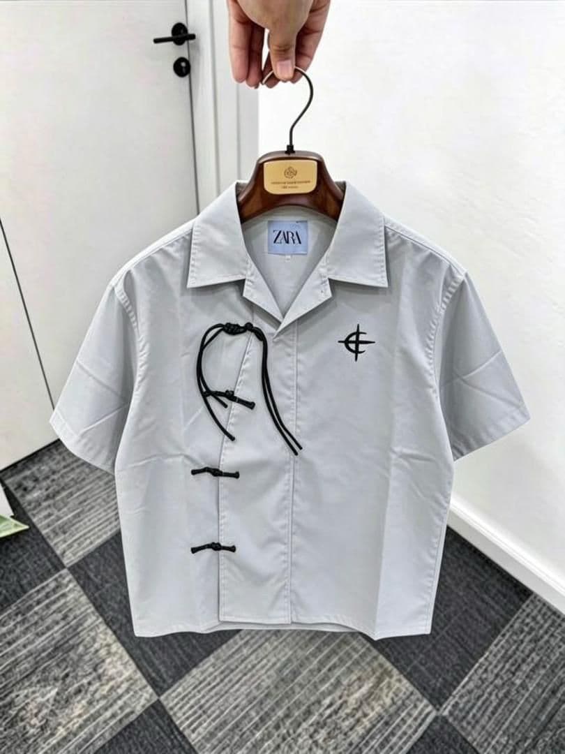 White short sleeve shirt with frog buttons, drawstring and chest embroidery on wooden hanger.